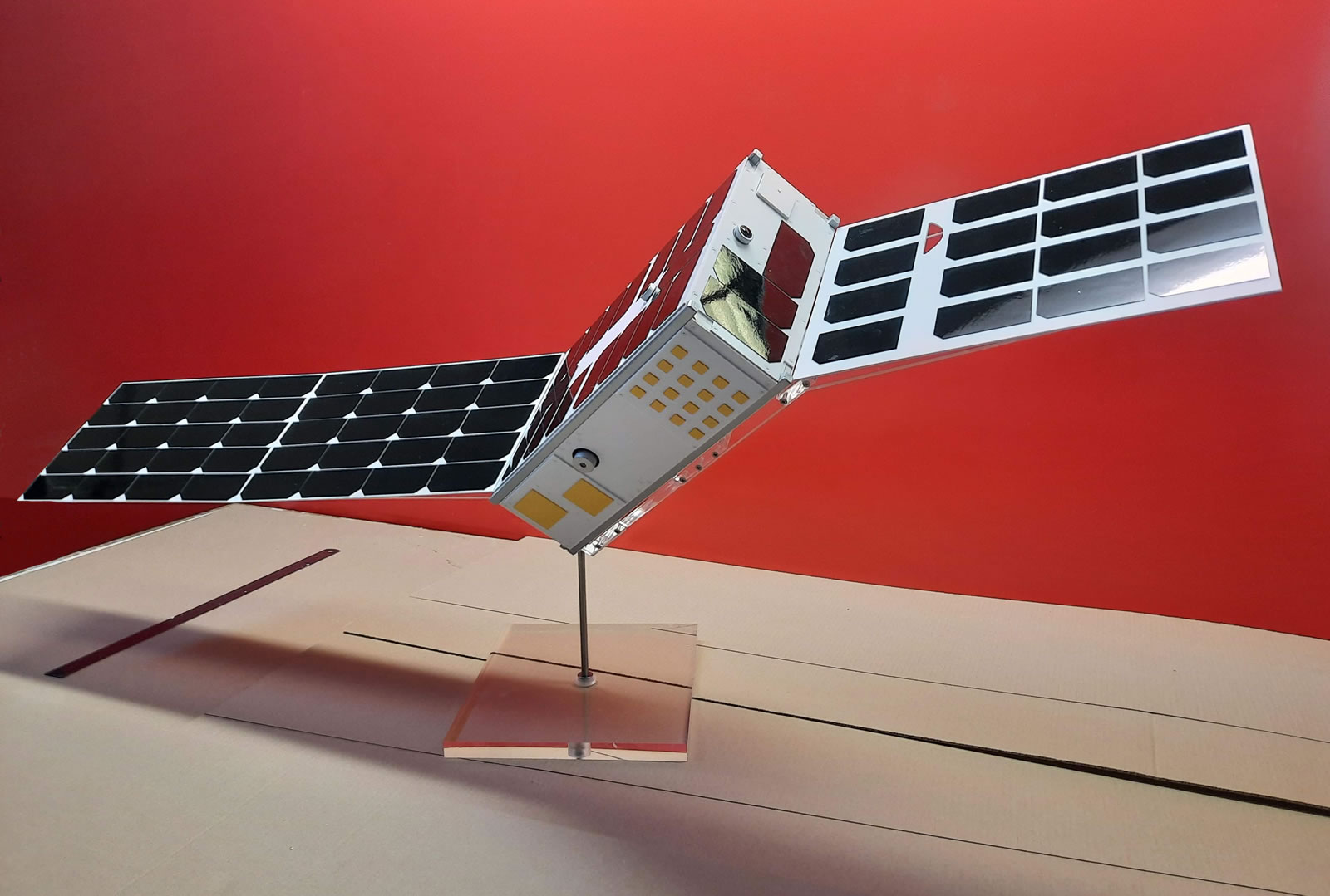 Maqueta de satélite aeroespacial con paneles solares laterales desplegados sobre soporte de exposición.